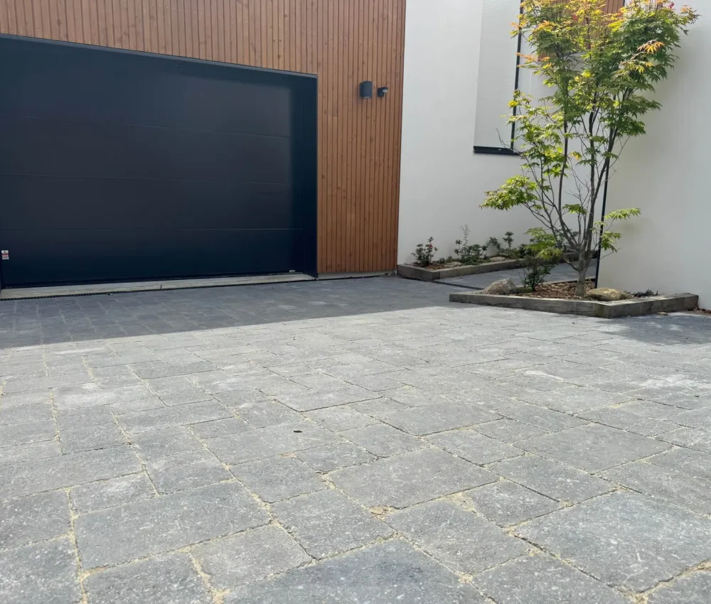 pavé de pavés gris en extérieur devant une porte de garage et un mur blanc avec un petit arbre